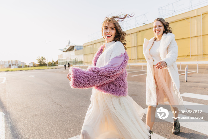 Charming laughing woman in glamorous attire walking down the street with friend. Amazing girls fooling around in warm october morning.