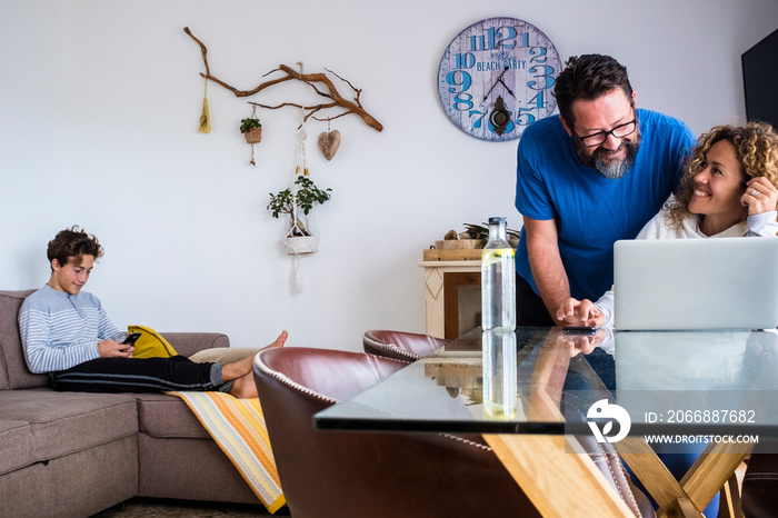 Caucasian family at home lifestyle with technology - couple husband and wife use laptop computer on the table and teenager son with phone on the couch - quarantine lockdown concept