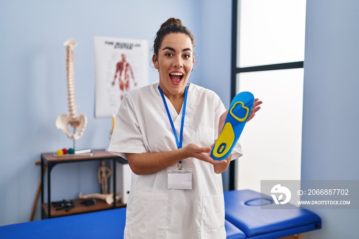 Young hispanic woman holding shoe insole at physiotherapy clinic celebrating crazy and amazed for success with open eyes screaming excited.