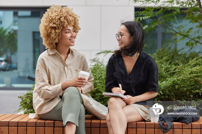 Diverse cheerful young women have pleasant conversation take interview write answers to questionnaire drinks takeaway coffee dressed in casual clothes pose on bench against urban background.