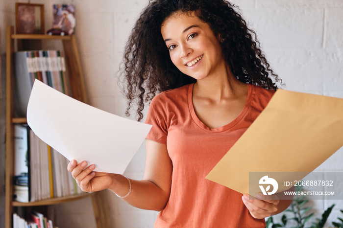 Young cheerful woman with dark curly hair in T-shirt joyfully looking in camera holding letter with exam results in hands at home