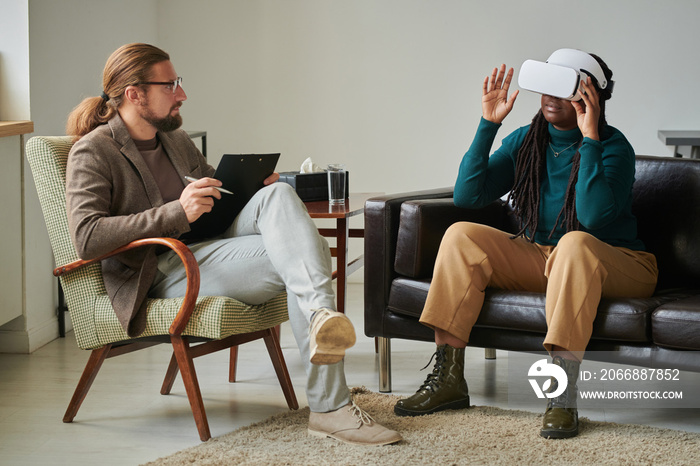 African American girl in wireless VR glasses sitting on sofa during psychotherapy with psychologist