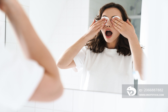 Photo of woman holding cotton pads on her eyes while looking at mirror