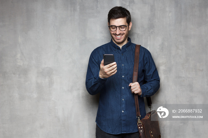 European modern smiling business man in glasses and shirt standing against textured wall with phone in hands and bag on shoulder