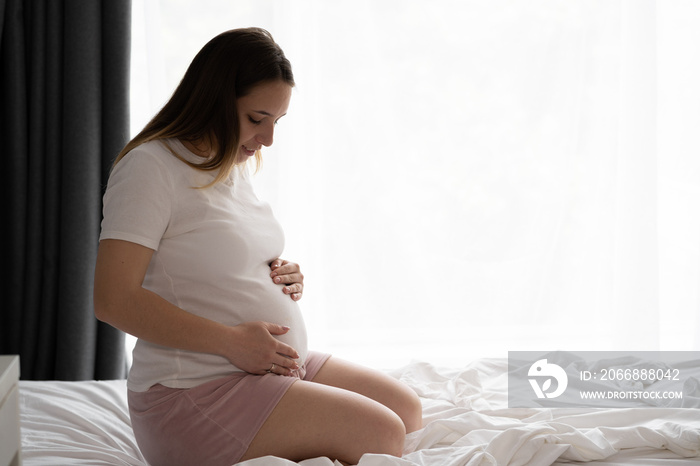 Attractive pregnant woman sitting in bed and holding her belly, bedroom window background. Last months of pregnancy.