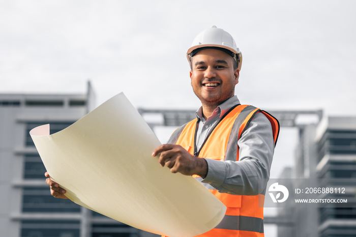 Asian engineer handsome man or architect open the paperwork blueprint and planning to building construction. Worker asian man with white safety helmet working in modern building construction site.