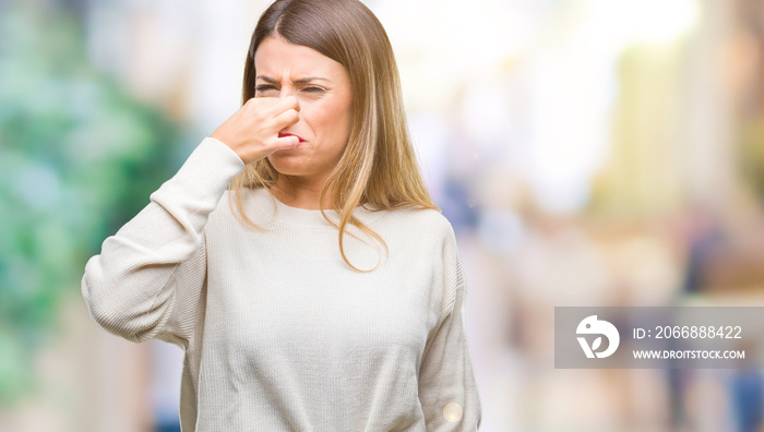 Young beautiful woman casual white sweater over isolated background smelling something stinky and disgusting, intolerable smell, holding breath with fingers on nose. Bad smells concept.