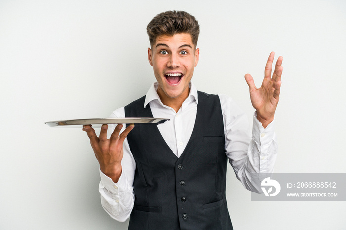 Young caucasian waitress man holding a tray isolated on white background receiving a pleasant surprise, excited and raising hands.