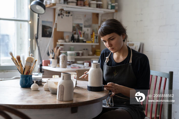 Young woman artist making white clay vasesits at table in workshop applying patterns with brush and paints. Attentive girl student works as apprentice for specialist in manufacture of ceramic dishes