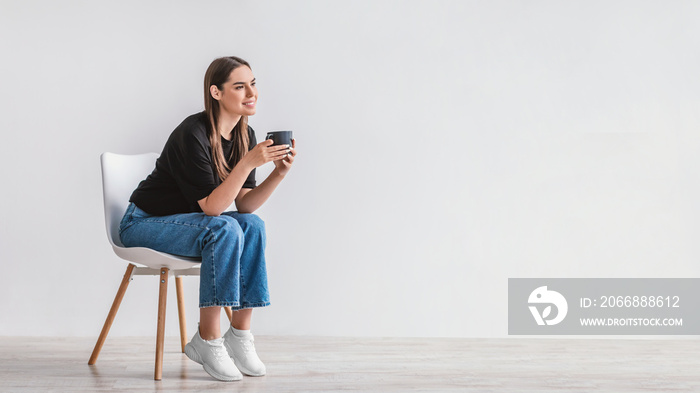 Coffee break. Dreamy young woman sitting on chair, enjoying cup of hot beverage against white wall, copy space