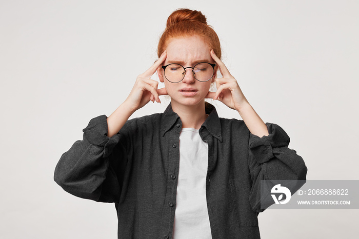 Portrait of a red-haired young girl student dressed in a black men’s shirt, which stands with her eyes closed, keeps her fingers on her temples, depicts a headache