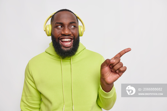 Horizontal shot of happy cheerful dark skinned hipster guy with thick beard listens music in headphones wears casual hoodie points away on blank space gives way or direction isolated on white