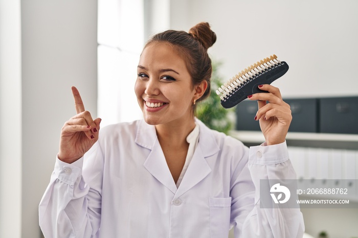 Young hispanic dentist woman holding teeth whitening samples surprised with an idea or question pointing finger with happy face, number one