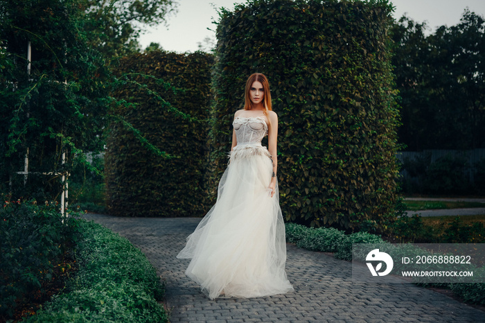 Young beautiful model is posing in a long ivory dress in the garden with a crown on her head