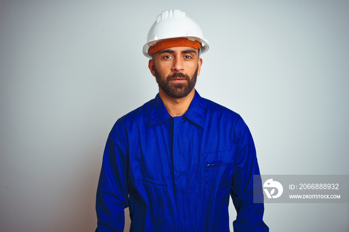 Handsome indian worker man wearing uniform and helmet over isolated white background Relaxed with serious expression on face. Simple and natural looking at the camera.