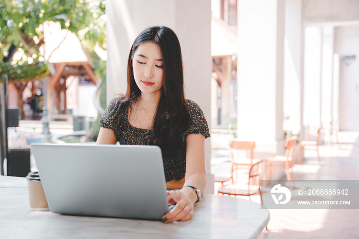 Beautiful Asian girl enjoys coffee while working in a coffee shop on laptop connected to public wifi. Stylish college student studying online while spending free time in the park.