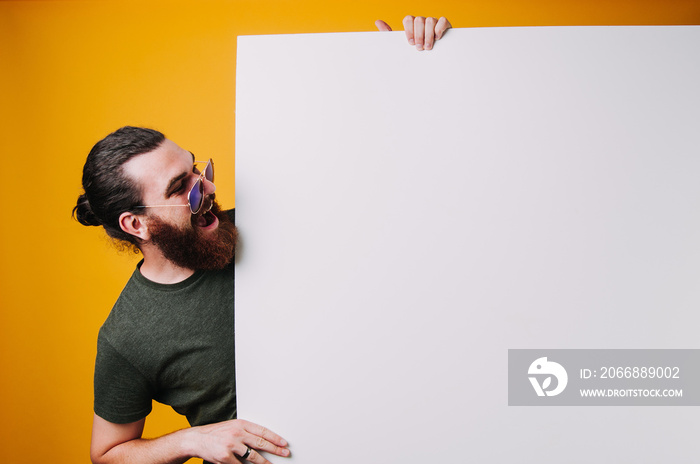 Hot young bearded man in gray with long hair and glasses amazed looking at your text on white sheet on yellow background.