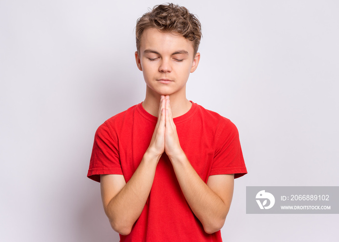 Portrait of teen boy praying, on grey background. Cute caucasian teenager with hands folded in prayer hoping for better. Child asking God for good luck, success or forgiveness.