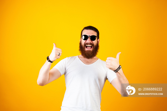 Excited young hipster bearded man wearing glasses and a white T-shirt laughing and smiling at the camera isolated on a yellow background. Thumbs up.