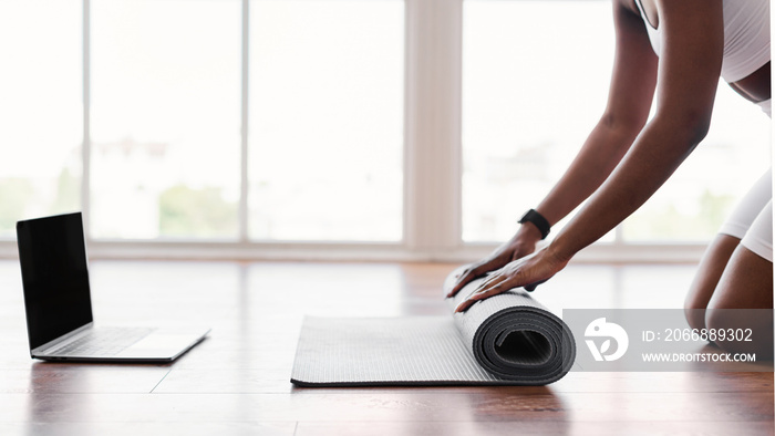 Young black woman unrolling yoga mat on floor near pc