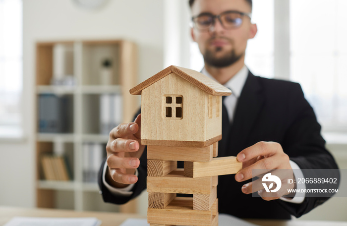 Male entrepreneur removes wooden blocks from tower foundation of little miniature toy house model as symbol of business risks and risky, dangerous, unstable situation on real estate market. Close up