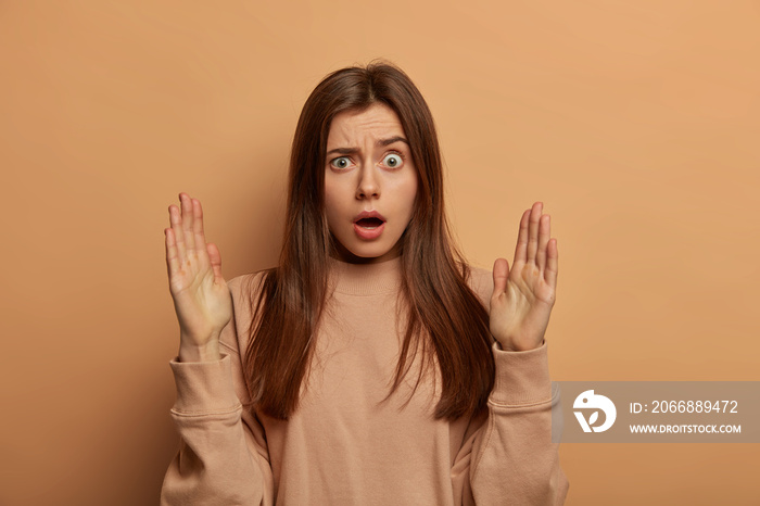 Impressed stupefied European woman shows something very big or huge, stares with bugged eyes, shapes object with both palms, wears casual sweatshirt, isolated over brown background. Size matters