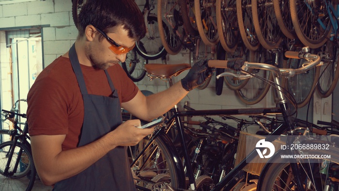 theme small business bike repair. Young caucasian brunette man wearing safety goggles, gloves and fartukhe uses mobile phone technology, takes notes, checklist in bicycle workshop