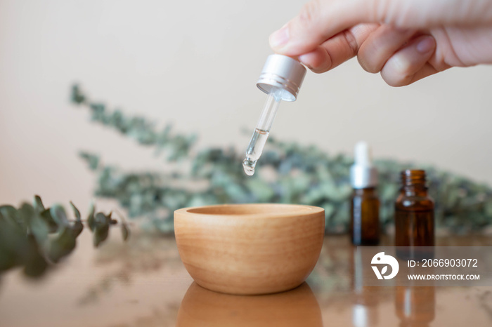 Woman’s hand holding pipette and dripping eucalyptus essential oil into a bowl with natural eucalyptus leaves on wooden table. Aromatherapy, spa, skincare or herbal medicine concept.