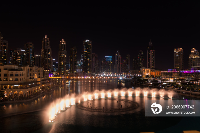 Dubai singing fountains at night lake view between skyscrapers. City skyline in dusk modern architecture in UAE capital downtown.