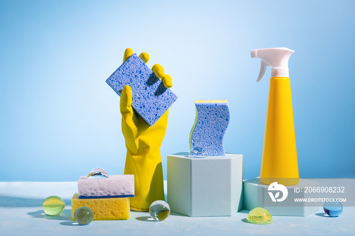 Creative still life with supplies for cleaning or housekeeping on podiums over blue background.Fenmale hand in yellow rubber glove with clean sponge, spray bottle and color sponges..