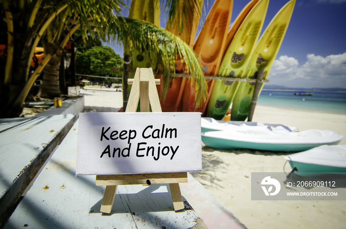 conceptual image with word KEEP CALM AND ENJOY on white canvas frame and wooden easel. Blurred image of beach and colorful kayaks background at sunny day.