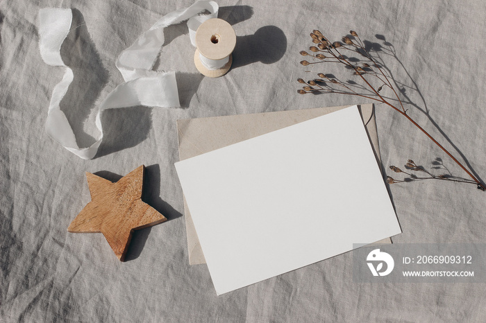 Christmas stationery. Blank greeting card, invitation mockup. Wooden star ornament, craft envelope and dry grass in sunlight. Beige linen tablecloth. White ribbon. Winter wedding flat lay, top