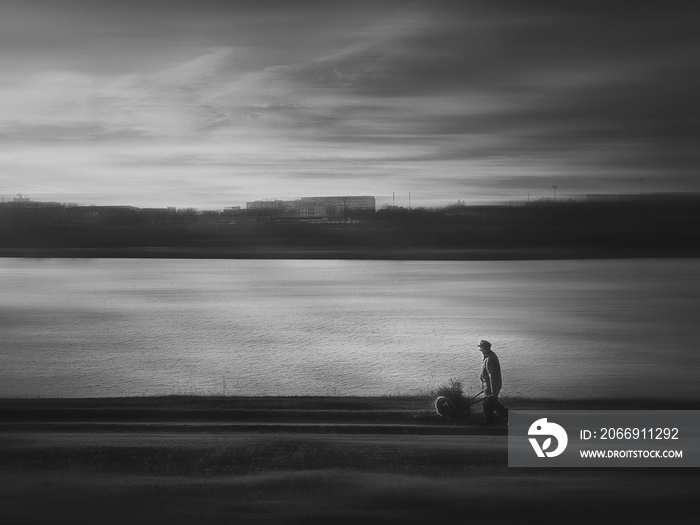 Moody countryside black and white scene, an old man carrying an antique trolley cart with firewood, going home, walking a dirt road track near the lake. Wanderer silhouette, silent evening atmosphere.