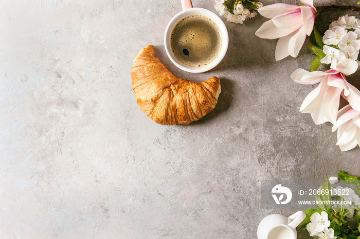 Pink mug of black espresso coffee, french croissant, cream and spring flowers magnolia, blooming cherry branches over grey texture background. Top view, space. Spring greeting card, wallpapers. Toned