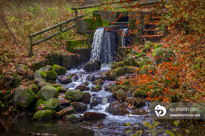 a small waterfall located in a forest surrounded by trees with colourful autumn leaves