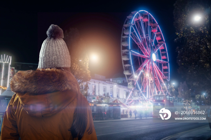 Teenager girl in focus, fun wheel and busy city street out of focus in the background. The model in warm winter clothes. Festive season celebrations concept. Light flare.