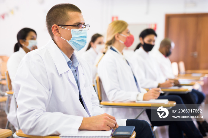 Focused man in white coat and protective mask sitting in conference room during medical seminar. Necessary precautions in coronavirus pandemic