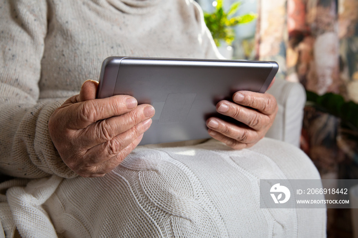 Close-up wrinkled hands of old woman with tablet computer. Senior lady sitting in chair with blanket. Warm and cozy, time to relax, browsing, watching movie.