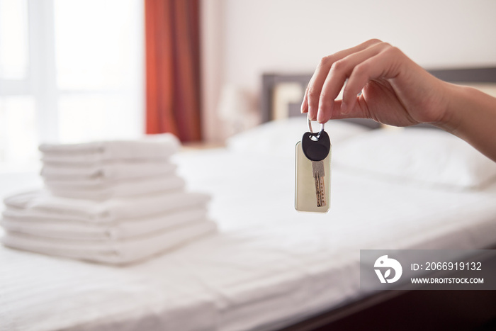 Woman holding in hand room key with blank label at hotel suite, copy space. Bedroom interior in blur