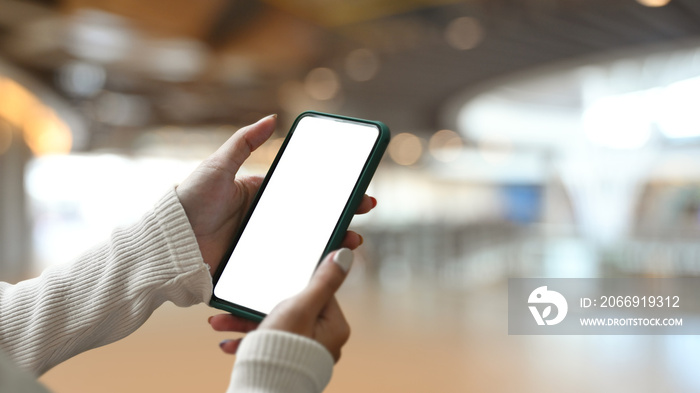 Close up view of young woman holding smart phone with empty screen on shopping mall blurred background.