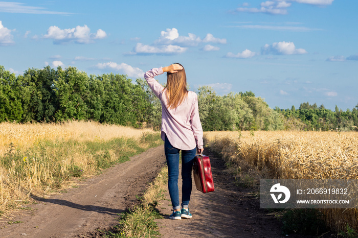 Beautiful young woman with a suitcase is walking along a road between wheat fields in the background. Travel concept, vacation