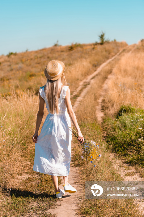 back view of young woman in white dress and straw hat holding wildflowers while walking on road in meadow