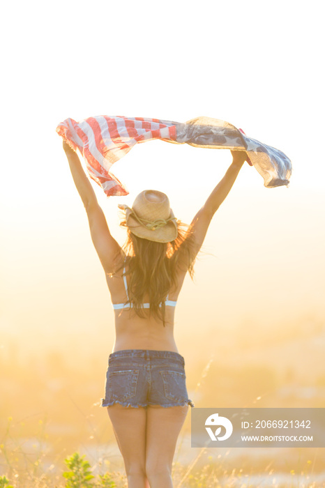 Woman wearing bra top and denim shorts, holding American flag above head, rear view