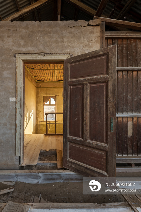 A vertical photograph inside an abandoned house, with an open door framing another room, taken in the ghost town of Kolmanskop, Namibia.