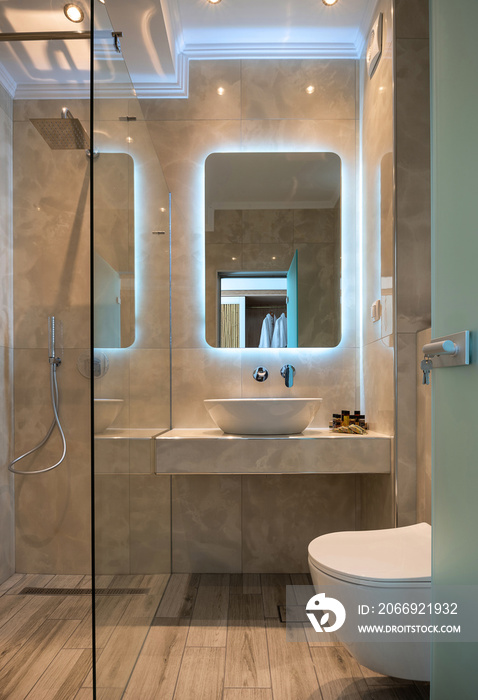 Front view of modern beige marble bathroom with vessel bowl sink, mirror back light, glass shower cabin, wall hung toilet, wooden floor