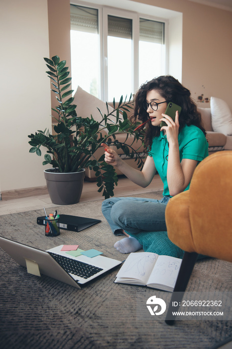 Lovely curly haired woman talking on phone while sitting on floor and doing homework with a computer and some books