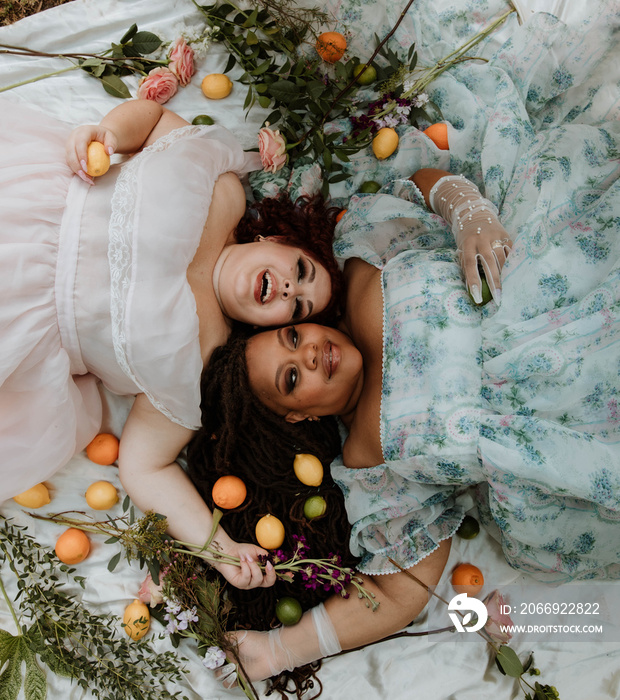 2 plus size women lie on the ground head to head holding fruit laughing