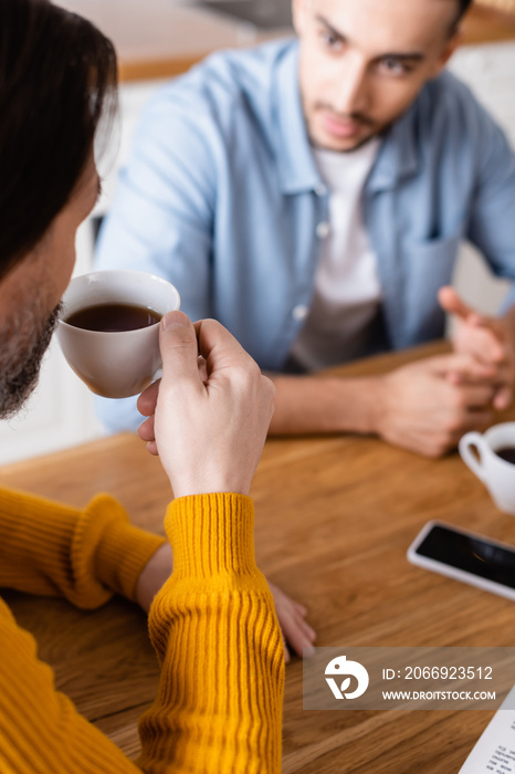 Man drinking coffee near young Hispanic son on blurred background in kitchen