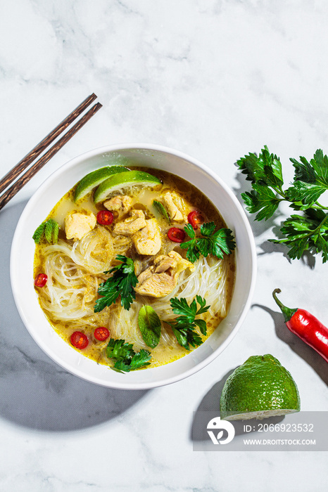 Thai green curry chicken soup with rice noodles, chili and herb in white bowl.