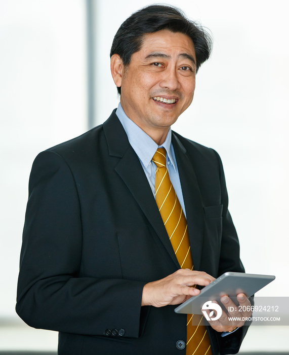 Portrait 50s Asian male executive visionary business male owner wearing formal suit and necktie, standing indoor office, using technology tablet, touching the screen, smiling confidently and reliably.